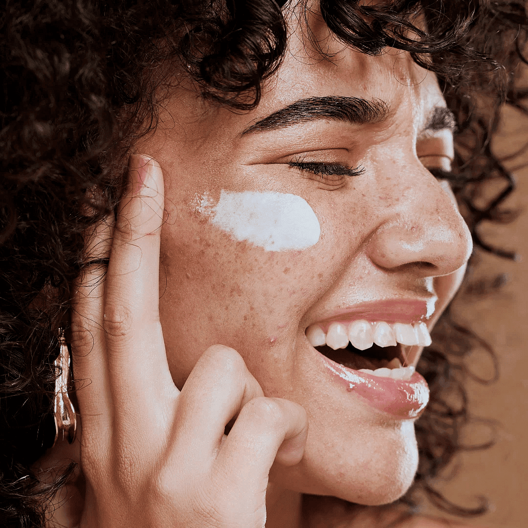 Woman with curly hair applying white cream to her face, smiling with eyes closed.